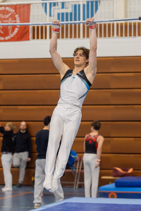 Young male gymnast hangs from the high bar with focused expression, wearing white and teal leotard in a gymnasium.