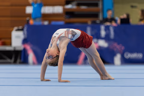 Young gymnast wearing glasses performs a bridge pose on the blue floor mat during an indoor gymnastics event.