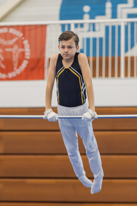 Young male gymnast hangs from the high bar with steady grip and calm, focused expression in a gymnasium.