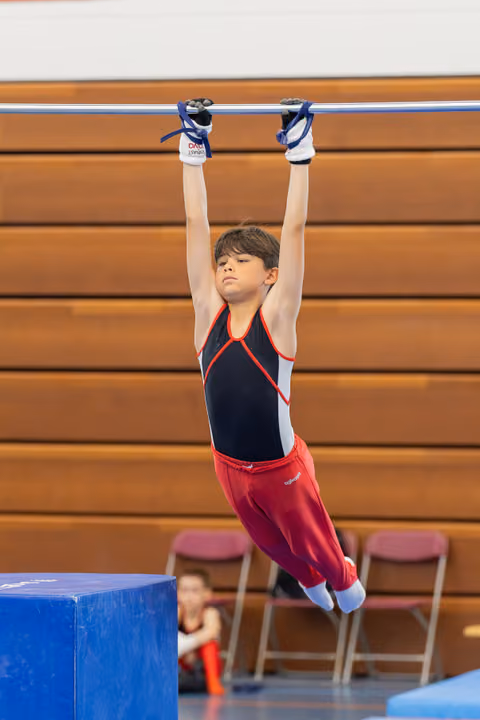 Young male gymnast hangs from the high bar with intense focus, wearing red and black gymnastics attire in an indoor gym.
