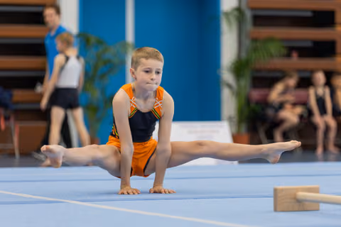 Focused young boy in orange leotard performs a straddle split on blue floor mat during gymnastics competition.