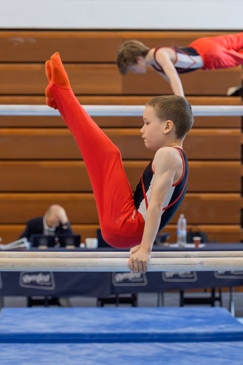 Young boy in red gymnastic suit holds a pike position on parallel bars, displaying intense focus and strength.
