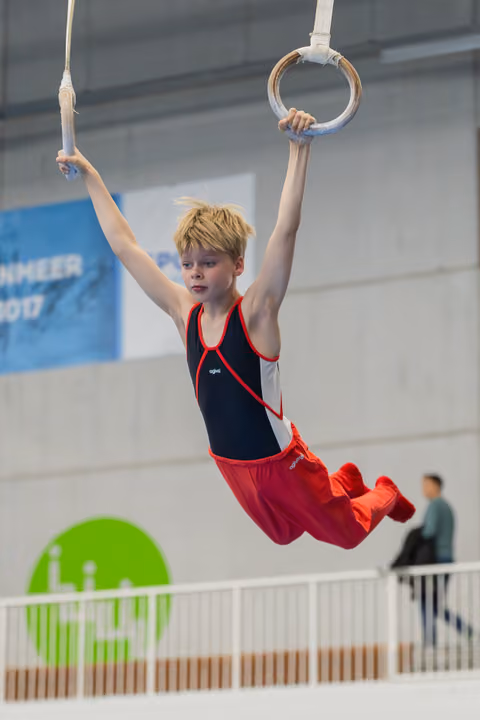 Blond boy hangs from gymnastic rings, legs tucked, focused expression, competing in an indoor gymnastics hall.