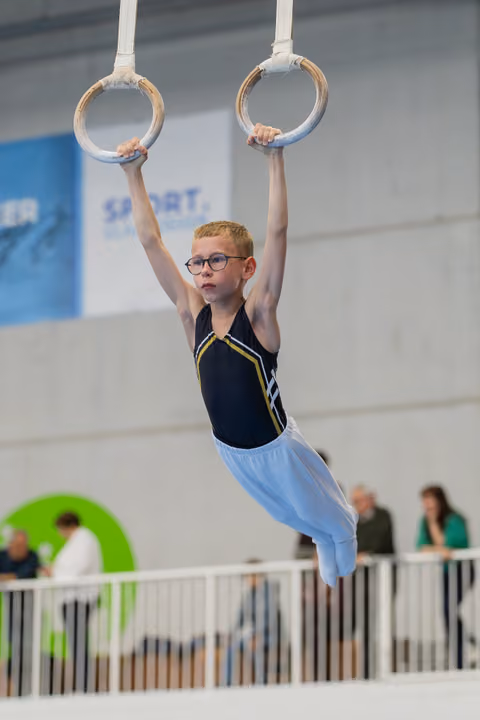 Young boy with glasses hangs with determination on gymnastic rings, body straight, in a sports hall during competition.