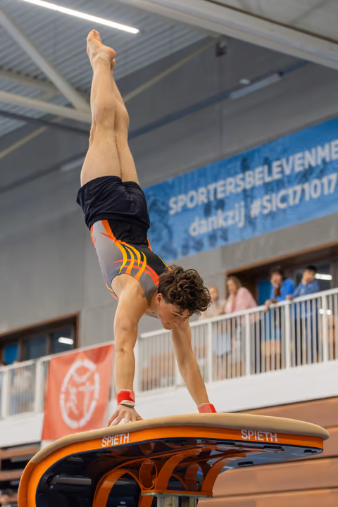 Young male gymnast performs a precise handstand on a Spieth vault, legs perfectly vertical, in an indoor sports hall.