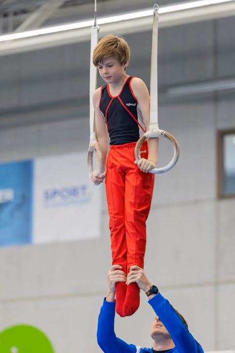 Young boy performs on gymnastics rings with intense focus while a coach in blue supports his feet below.