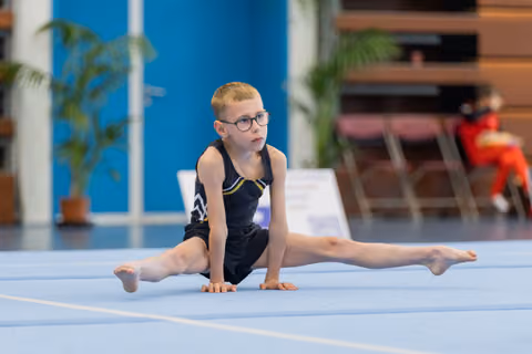 Young boy with glasses performs splits on blue gymnastics mat, focused expression, wearing black leotard during competition.