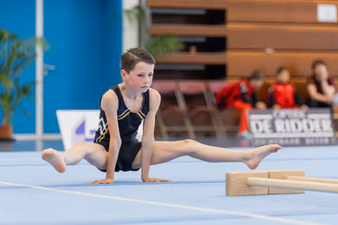 Focused young boy holds a straddle support position on the blue floor mat during a gymnastics competition.