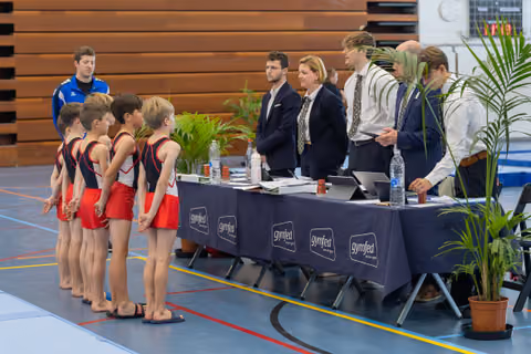 Young male gymnasts in red-black uniforms stand at attention before Gymfed judges at an official scoring table indoors.