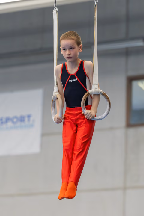 Young boy holds gymnastic rings with steady grip and focused expression, wearing red and black leotard in sports hall.
