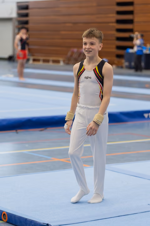 Young male gymnast smiles confidently on the floor mat, wearing white leotard and wrist guards at an indoor gymnastics meet.