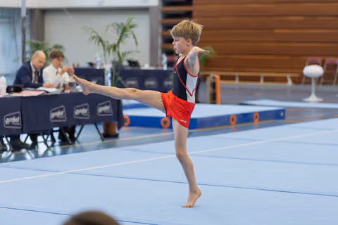 Young male gymnast performs a standing leg raise on the floor exercise mat, focused and precise, with judges scoring in background.