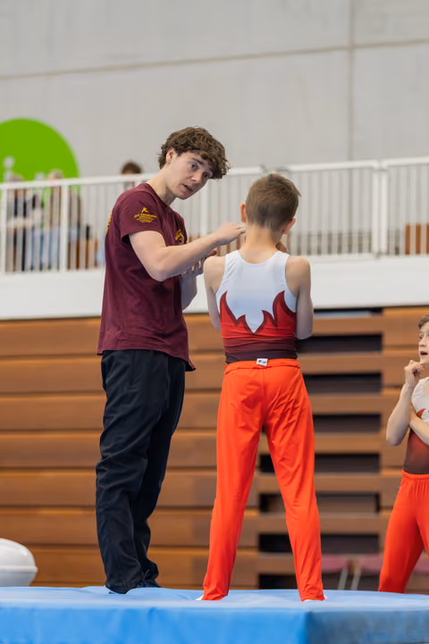 A teenage coach attentively gives feedback to a young male gymnast in a red and white leotard at an indoor gymnastics event.