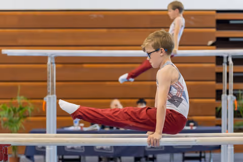 Focused young gymnast with glasses performs a straddle hold on parallel bars, legs extended forward, intense concentration.