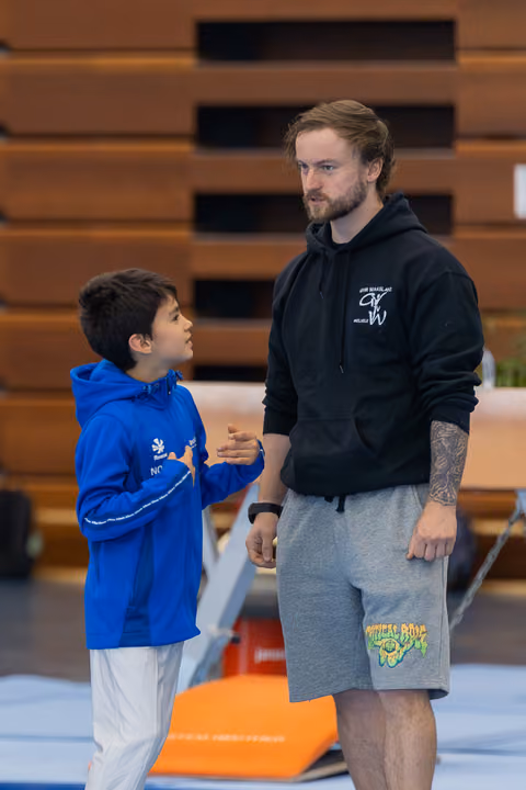 A young gymnast in a blue hoodie speaks earnestly to his coach, who listens attentively in a gymnasium setting.