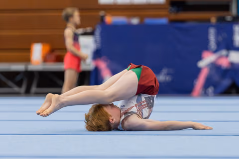 Young gymnast performs a floor routine, legs raised in a pike position, red-and-white leotard, indoor gymnastics hall.