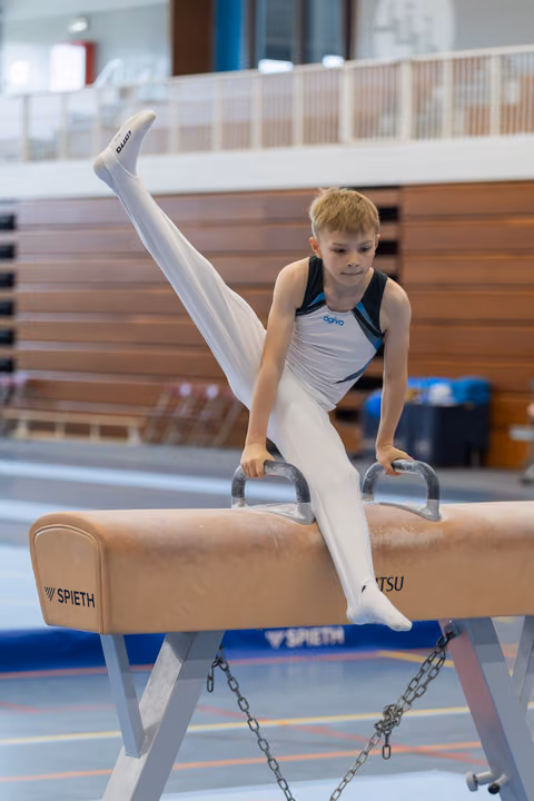 Focused young boy performs a leg split on the pommel horse, gripping the handles with determination in a bright gymnasium.