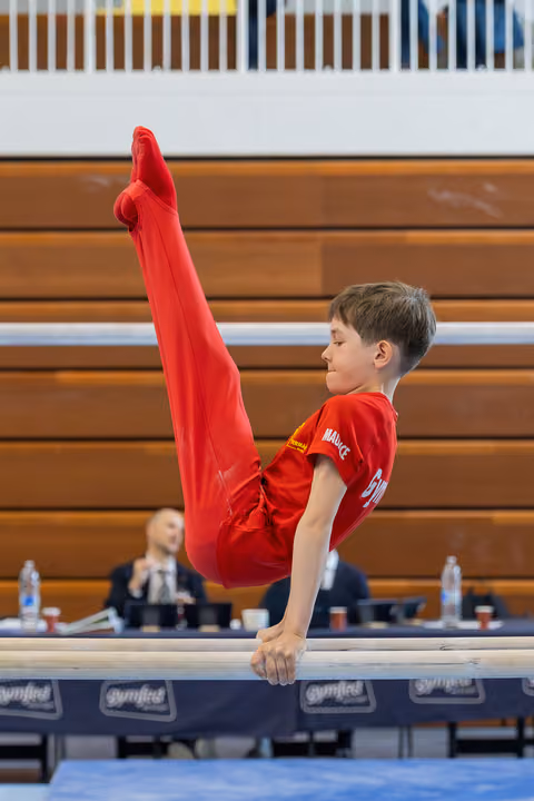 Young boy in red performs a straddle hold on parallel bars, face calm and focused during a gymnastics meet.