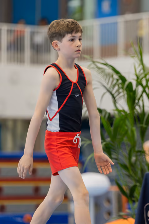 Young male gymnast walks with focused expression, wearing black and red leotard, in an indoor gymnastics venue.