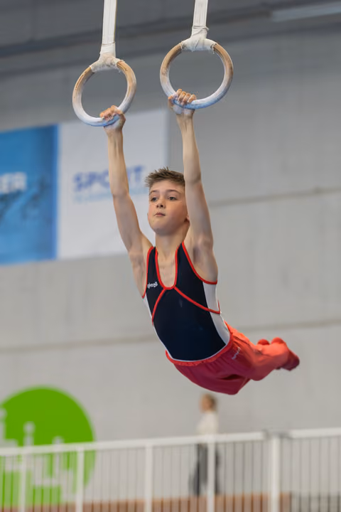 Young male gymnast hangs focused on still rings, body arched, wearing navy and red leotard in an indoor gym.