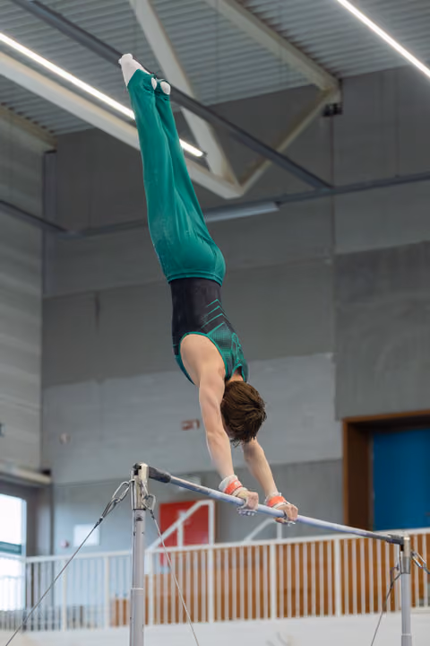 Male gymnast holds a perfect handstand on the high bar, body fully extended upside down in a teal competition leotard.