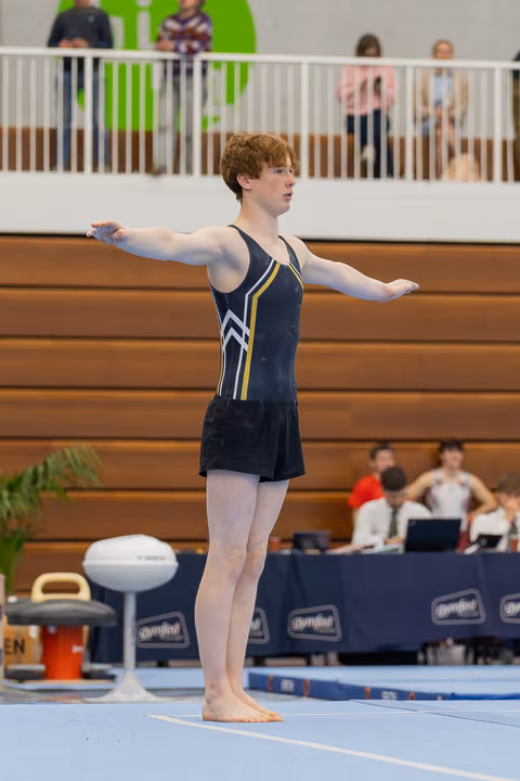 Red-haired teenage gymnast stands poised on the floor mat, arms outstretched with focused concentration during his routine.