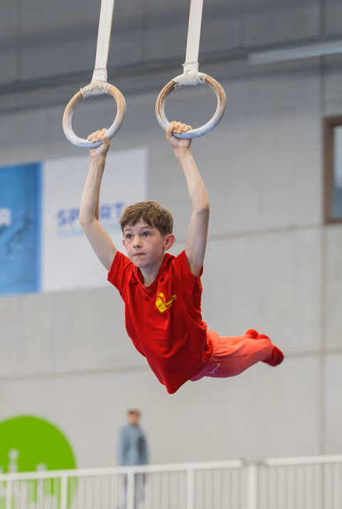 Focused young boy in red kit hangs from gymnastic rings mid-routine at an indoor sports hall competition.