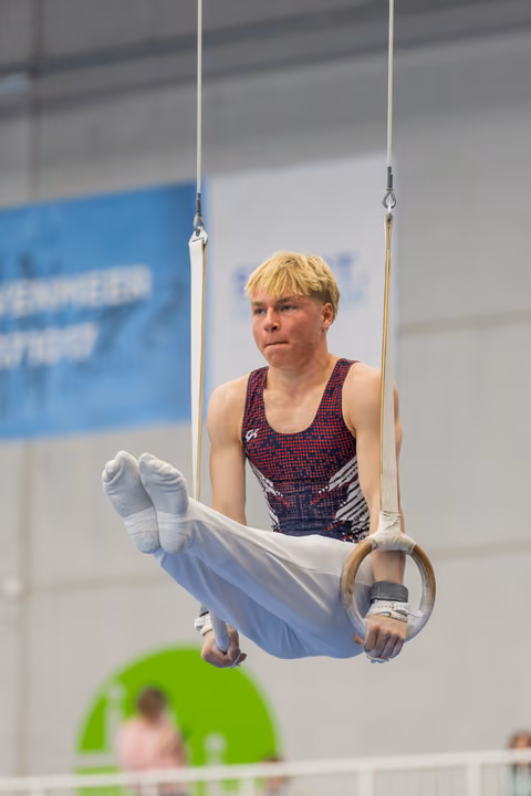 Young male gymnast grips still rings with intense concentration, legs tucked, competing in an indoor gymnastics hall.