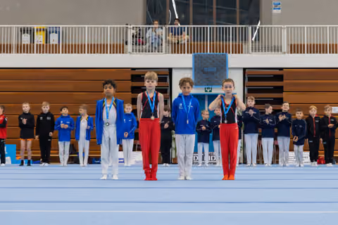 Four young gymnasts stand proudly on the floor mat, displaying their medals at an indoor gymnastics award ceremony.