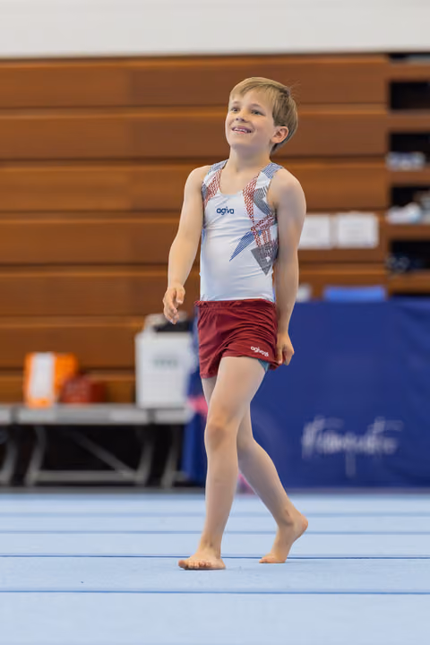 Young boy in agiva gymnastics attire walks barefoot on floor mat, smiling proudly during a gymnastics event.