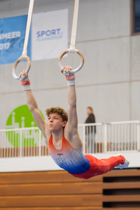 Young male gymnast hangs with intense focus on still rings, body extended horizontally in a dramatic hold.