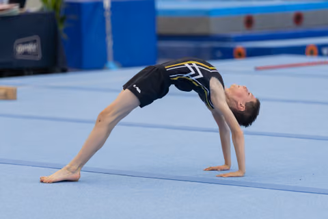 Young male gymnast holds a back bridge position on blue floor mat, body arched, focused and controlled.
