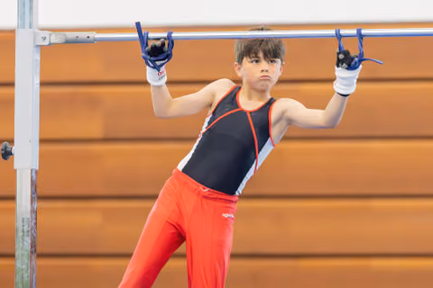 Young male gymnast grips the high bar with intense focus, wearing a black and red leotard with red pants.