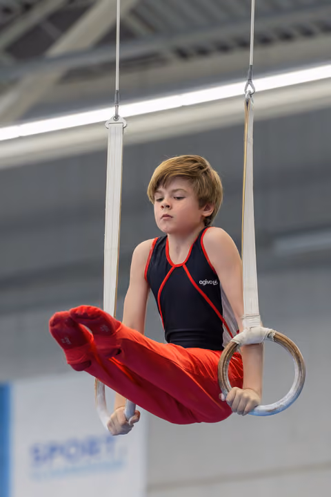 Focused young boy grips still rings mid-routine, legs tucked, wearing black and red gymnastic suit indoors.