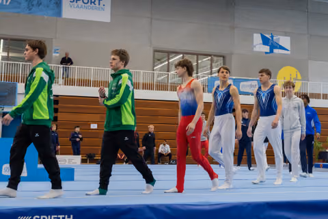 Male gymnasts in green and blue leotards walk across the floor mat during a gymnastics meet in Belgium.