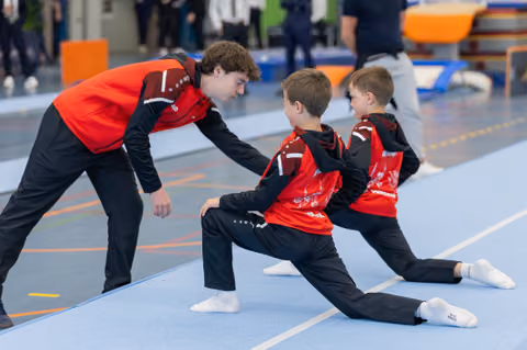 A teenage coach attentively guides two young boys doing splits stretches on a blue gymnastics mat, all in red uniforms.