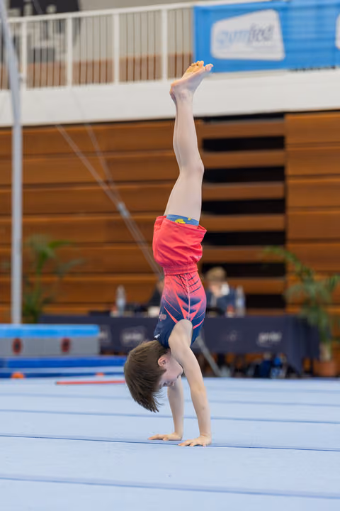 Young boy performs a straight handstand on the floor exercise mat during a gymnastics competition, legs extended upward.
