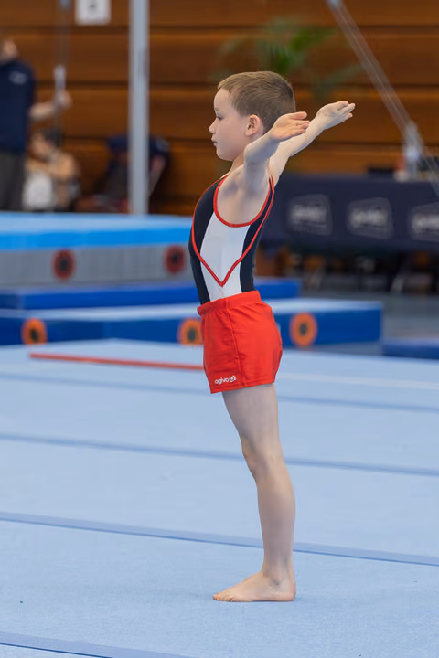 Young male gymnast stands tall on the floor mat, arm raised in a proud opening salute during a gymnastics meet.