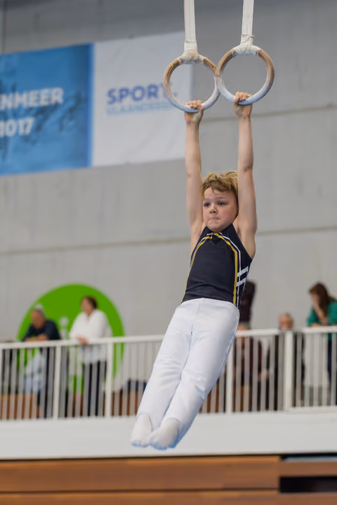 Determined young gymnast hangs from still rings, legs together, intense focus on his face during an indoor competition.