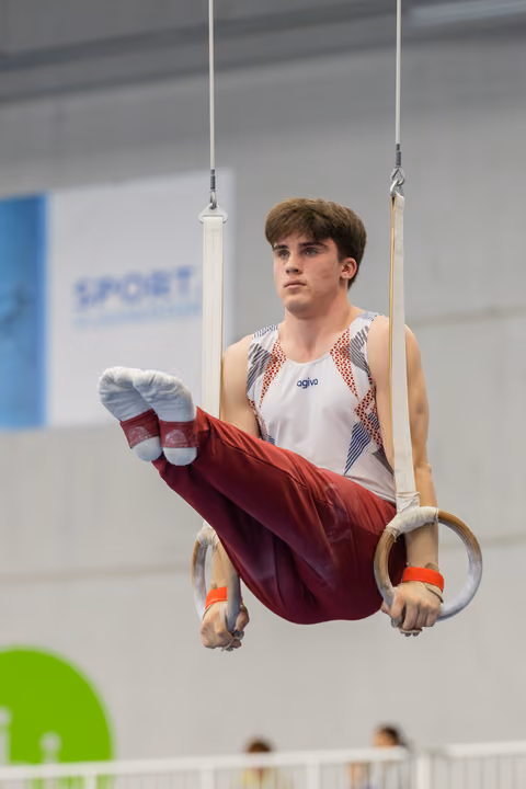 Young male gymnast holds a tuck position on still rings, focused expression, wearing agiva kit in a sports hall.