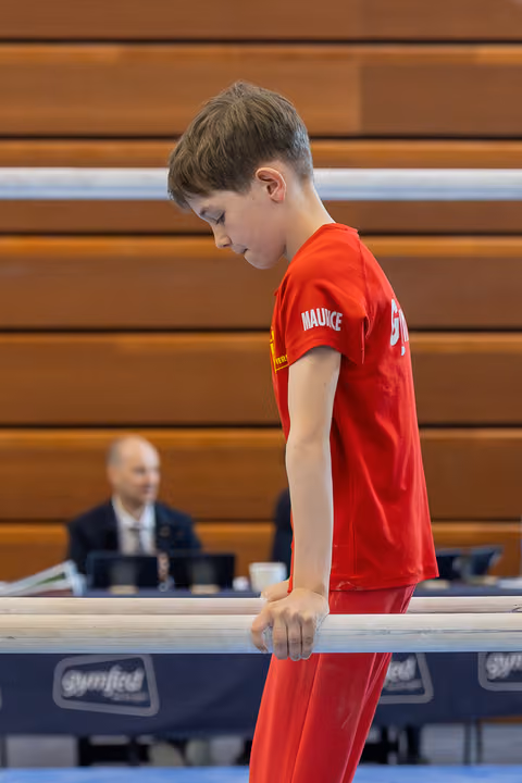 Boy named Maurice in red looks down with focus, gripping parallel bars at a gymnastics competition.