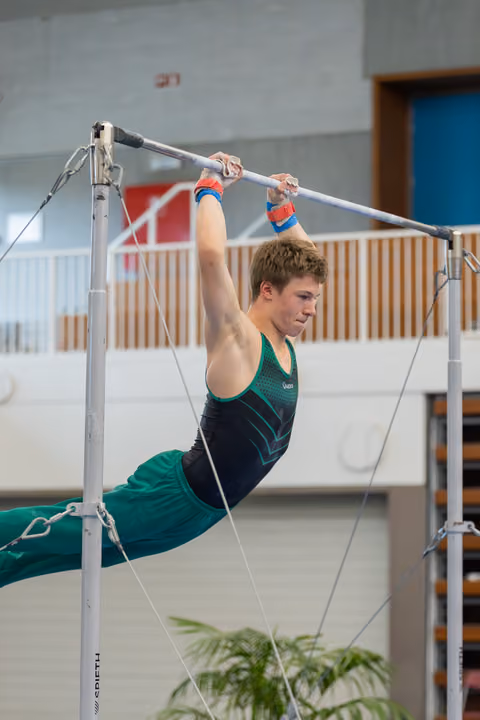 Young male gymnast hangs in a pike position on the high bar, face set with fierce concentration during indoor competition.