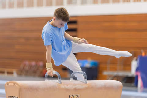 Young boy in blue tee concentrates intensely while performing on a Fujitsu pommel horse, leg raised mid-routine.