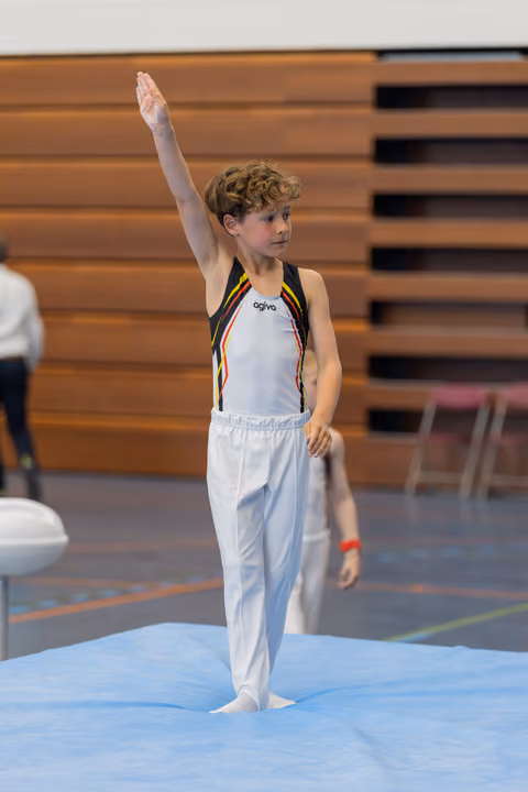 Young male gymnast raises one arm to salute the judges, standing barefoot on the floor exercise mat, composed and focused.