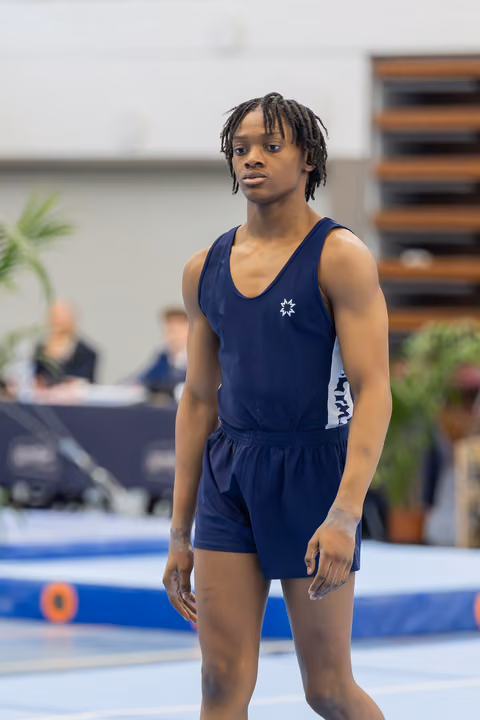 Young male gymnast in navy leotard stands composed and focused on the floor exercise mat, ready to perform.