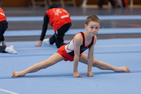 Young boy in red gymnastic suit performs a splits stretch on blue mat, looking focused and confident.