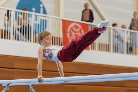 Focused young male gymnast executes a straddle L-sit on parallel bars before a crowd in a gymnasium.