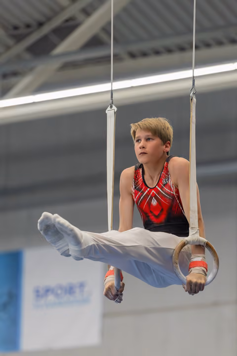 Young male gymnast holds a L-sit on still rings, focused gaze upward, wearing red and black leotard in an indoor gym.