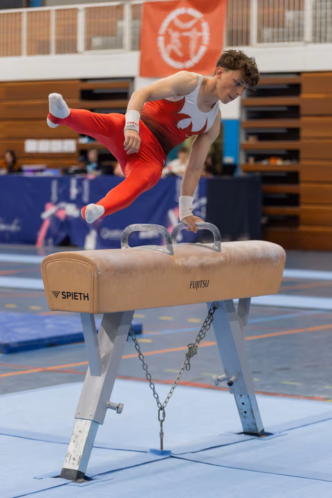 Young male gymnast in red suit performs dynamic move on pommel horse, legs extended mid-air with focused expression.