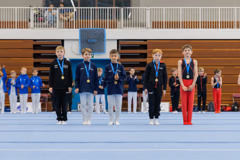 Five young male gymnasts stand proudly on the floor wearing gold medals, lined up at an indoor gymnastics competition.